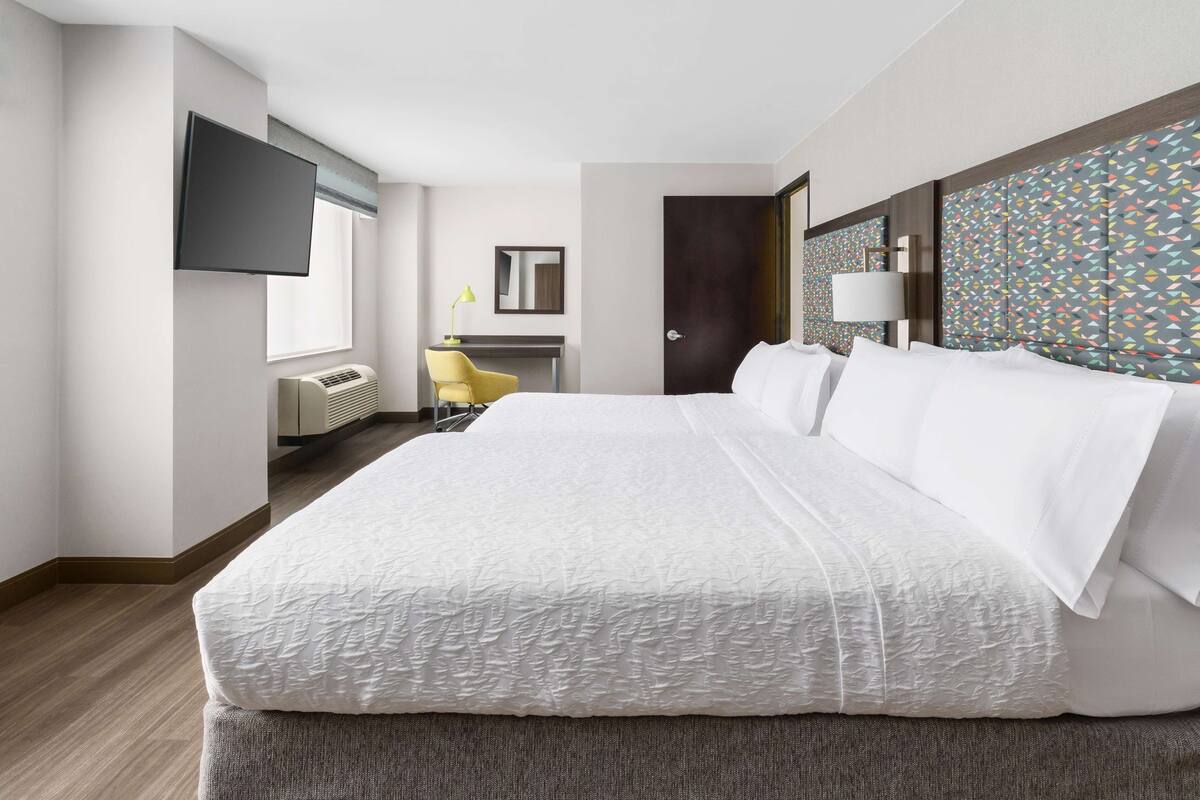 A hotel room with a large bed, a desk with a chair, a flat-screen TV mounted on the wall, and a patterned headboard.