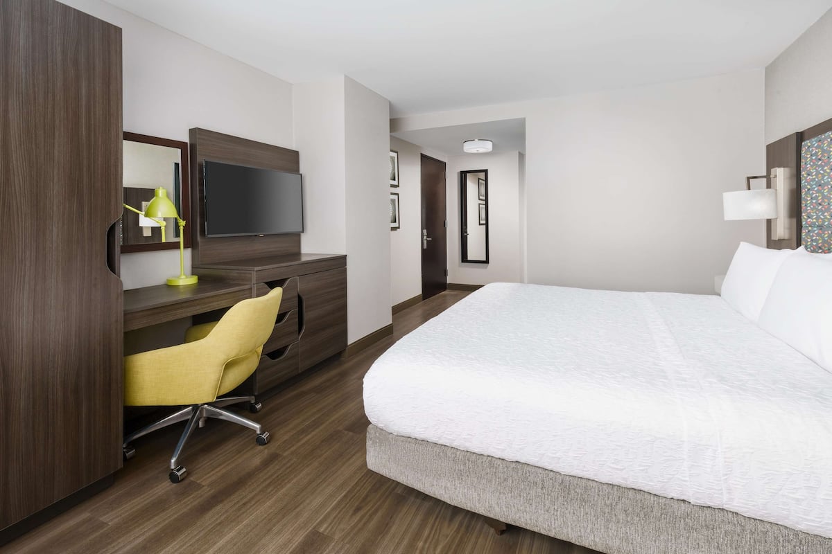 A hotel room with a large bed, a desk with a chair, a television, and a door leading to another room.