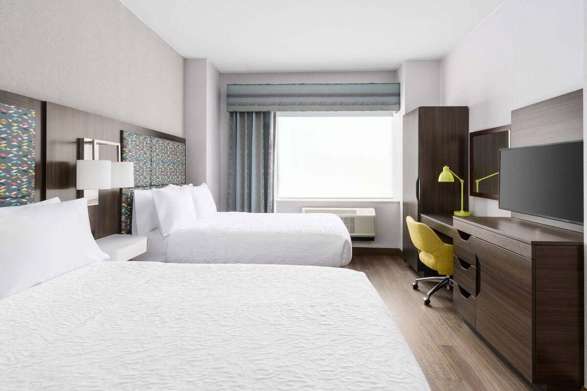 A hotel room with two beds, a desk with a TV, a chair, and a large window with curtains.