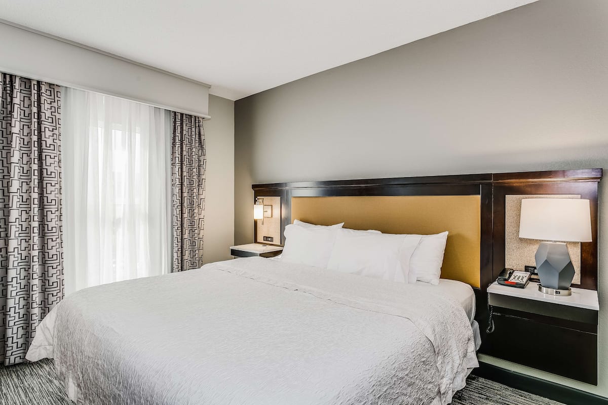A hotel room with a large bed, a nightstand with a lamp, and a window with patterned curtains.