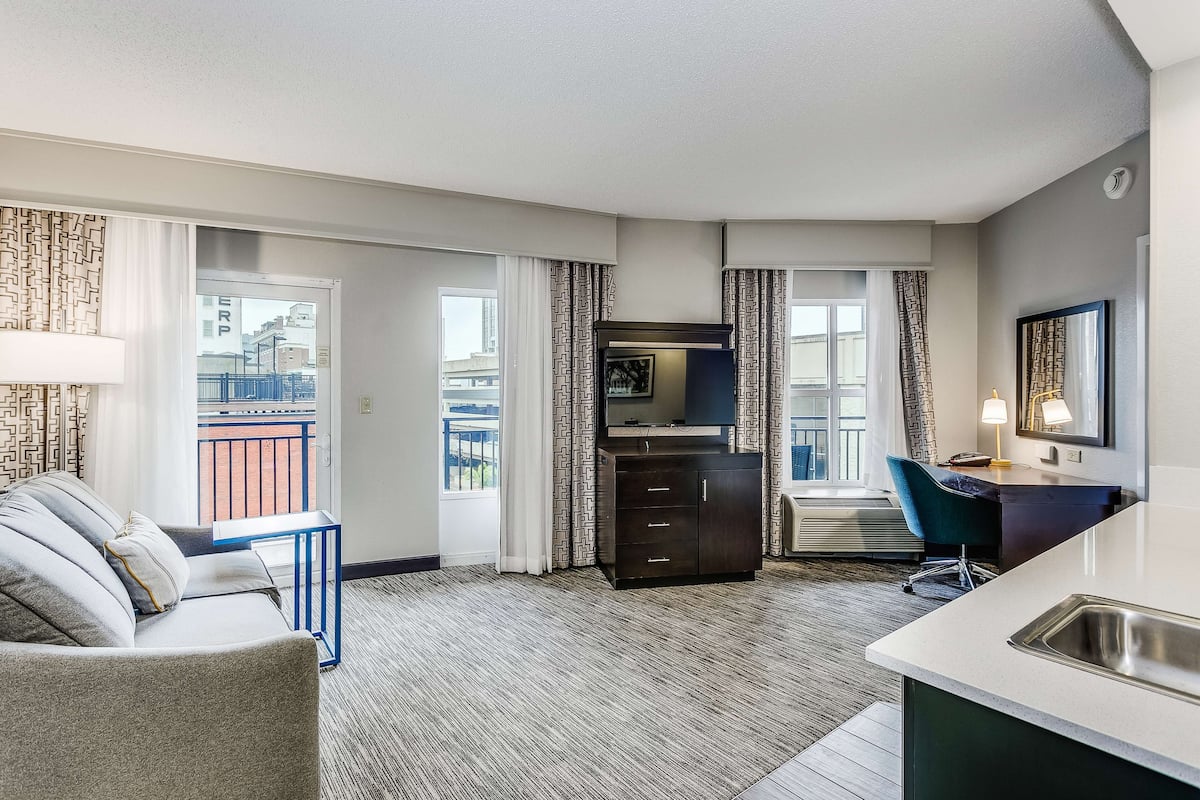 A modern hotel room with a sofa, a desk, a small kitchenette, and a balcony with a view of the city.