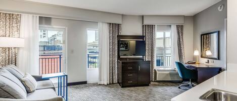 A modern hotel room with a sofa, a desk, a small kitchenette, and a balcony with a view of the city.
