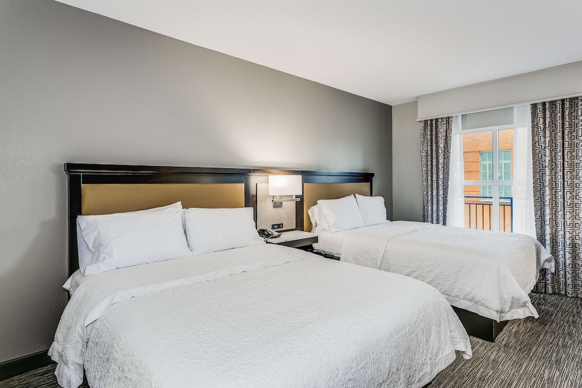 A hotel room with two beds, a nightstand, a lamp, and a window with curtains.