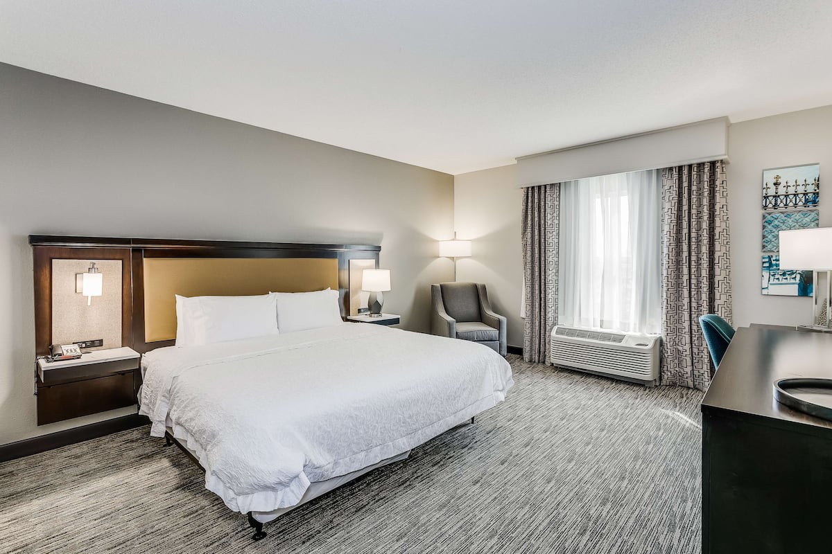 A hotel room with a large bed, a desk, a chair, a nightstand, a lamp, and a window with curtains.