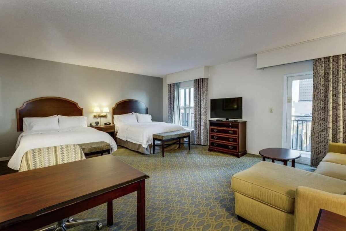 A hotel room with two beds, a TV, a desk, a chair, and a balcony with patterned curtains.