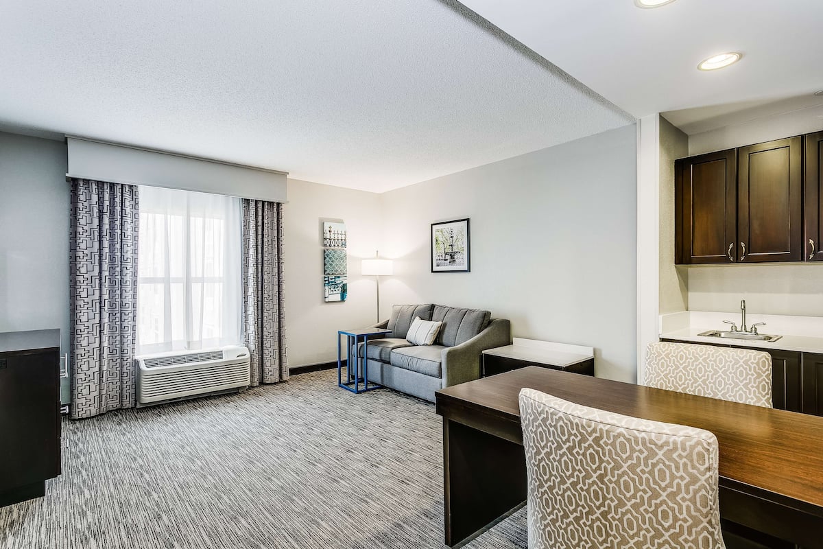 A hotel room with a sofa, a dining table, a lamp, a window with curtains, a kitchenette, and a wall-mounted picture.