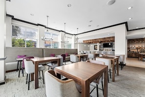 A modern dining area with high tables and chairs, a bar area with wooden cabinets, and large windows providing natural light.