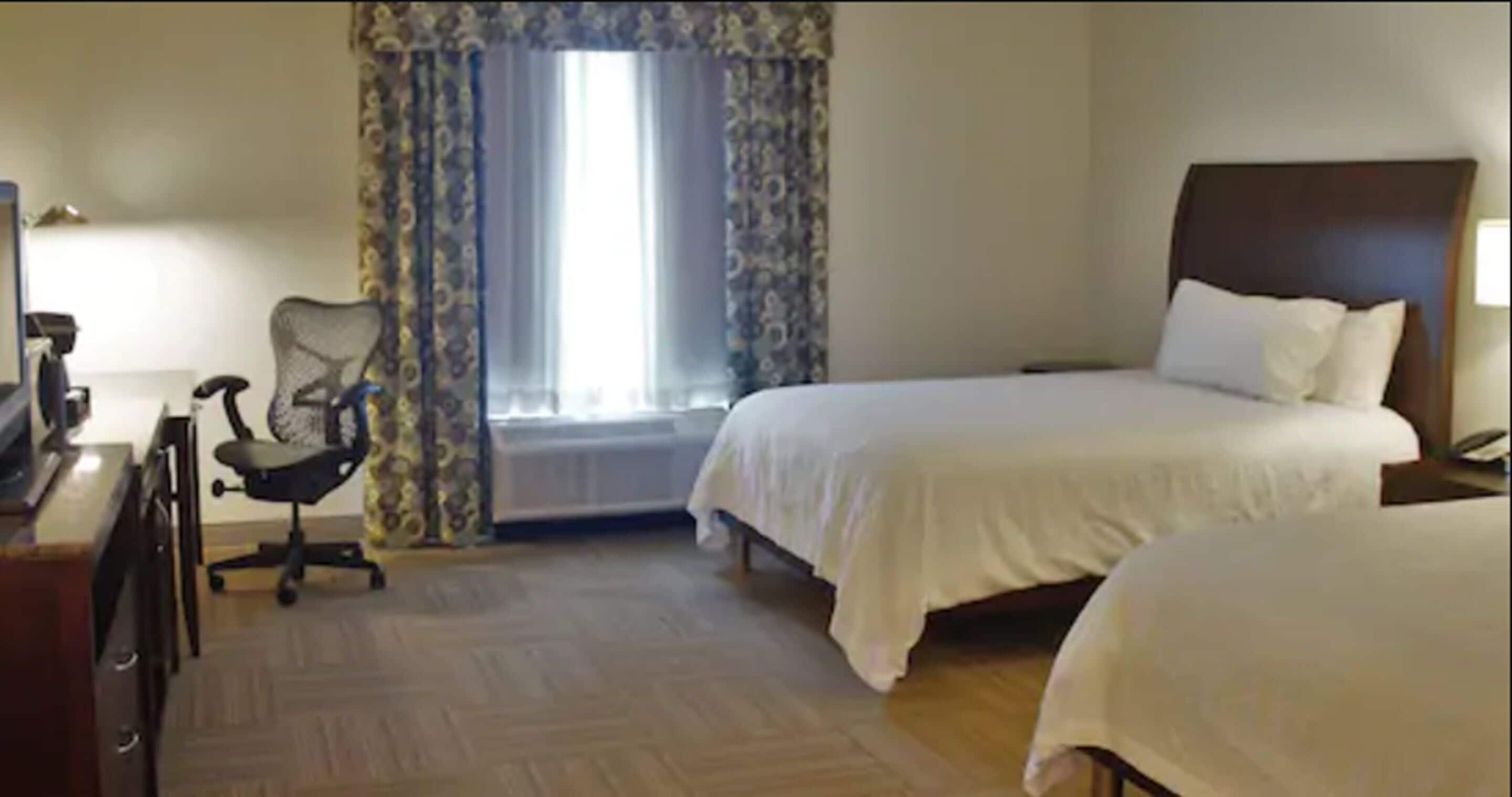 A hotel room with two beds, a desk with a chair, a window with curtains, and a radiator.