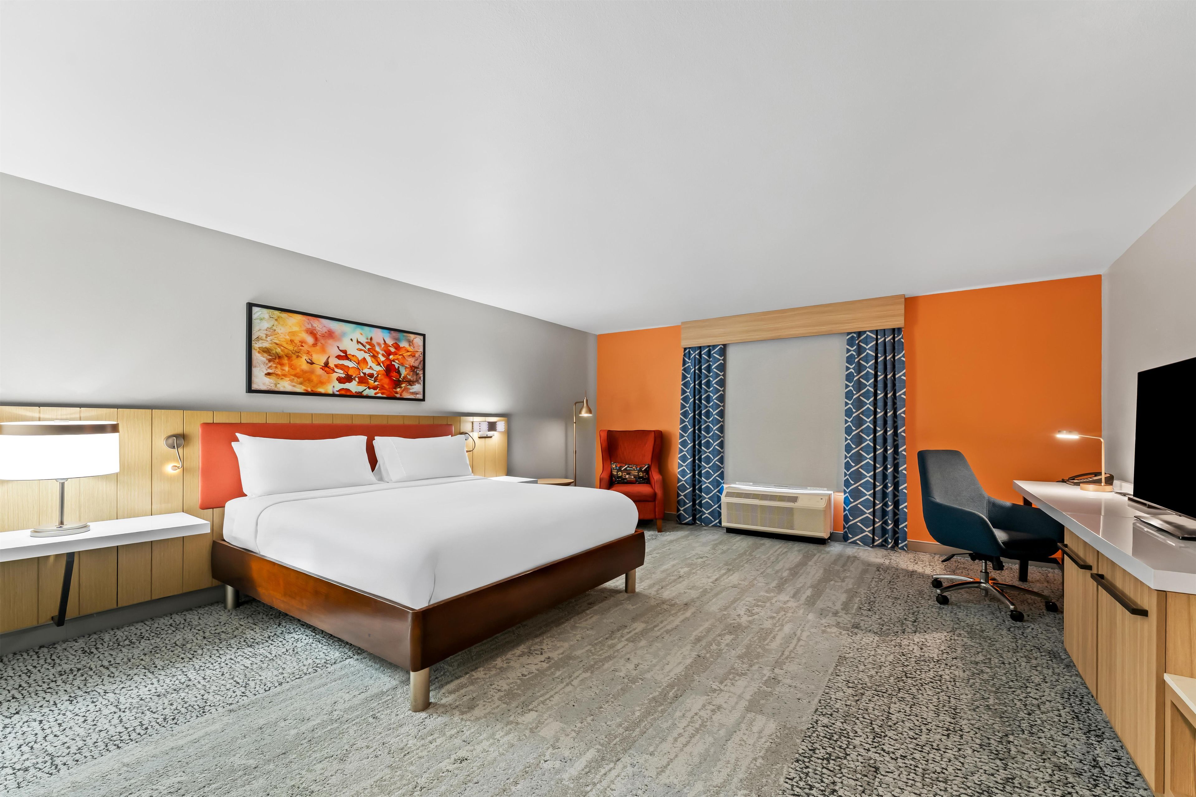 A hotel room with a bed, desk, chair, and a large window with curtains.