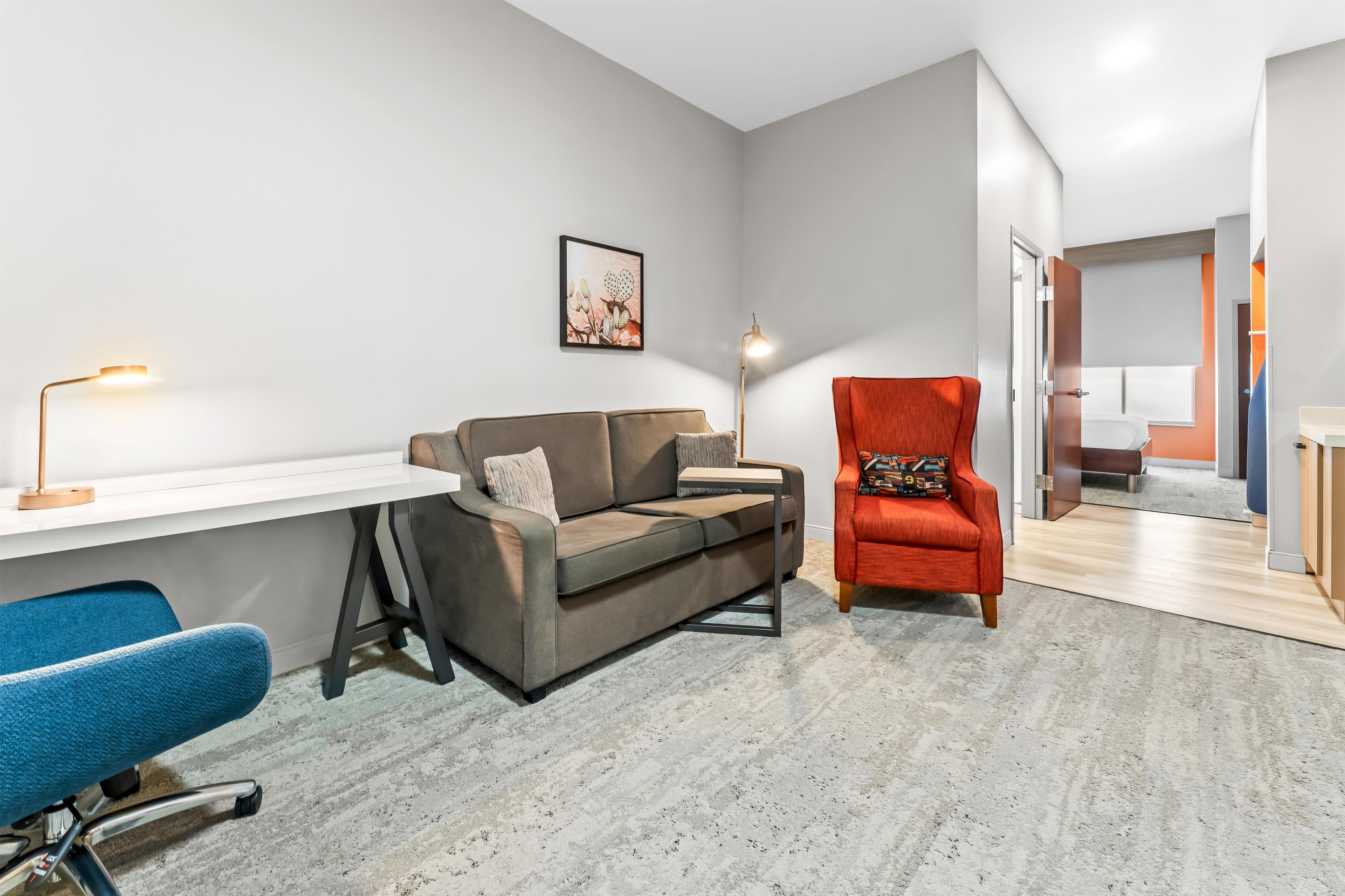 A modern living room with a white desk, a grey sofa, a blue chair, and a red armchair.