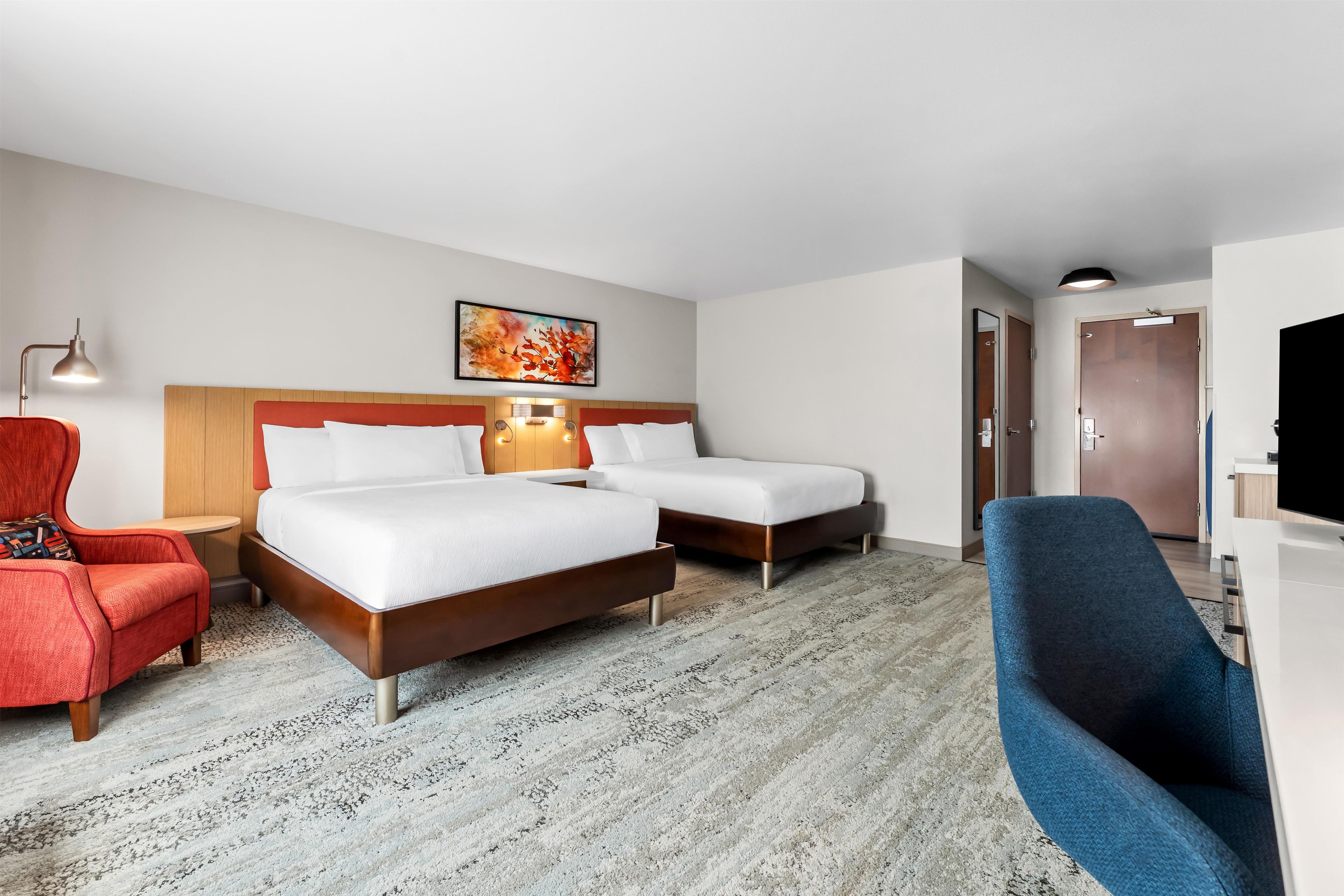 A hotel room with two beds, a desk, a chair, a lamp, and a painting on the wall.