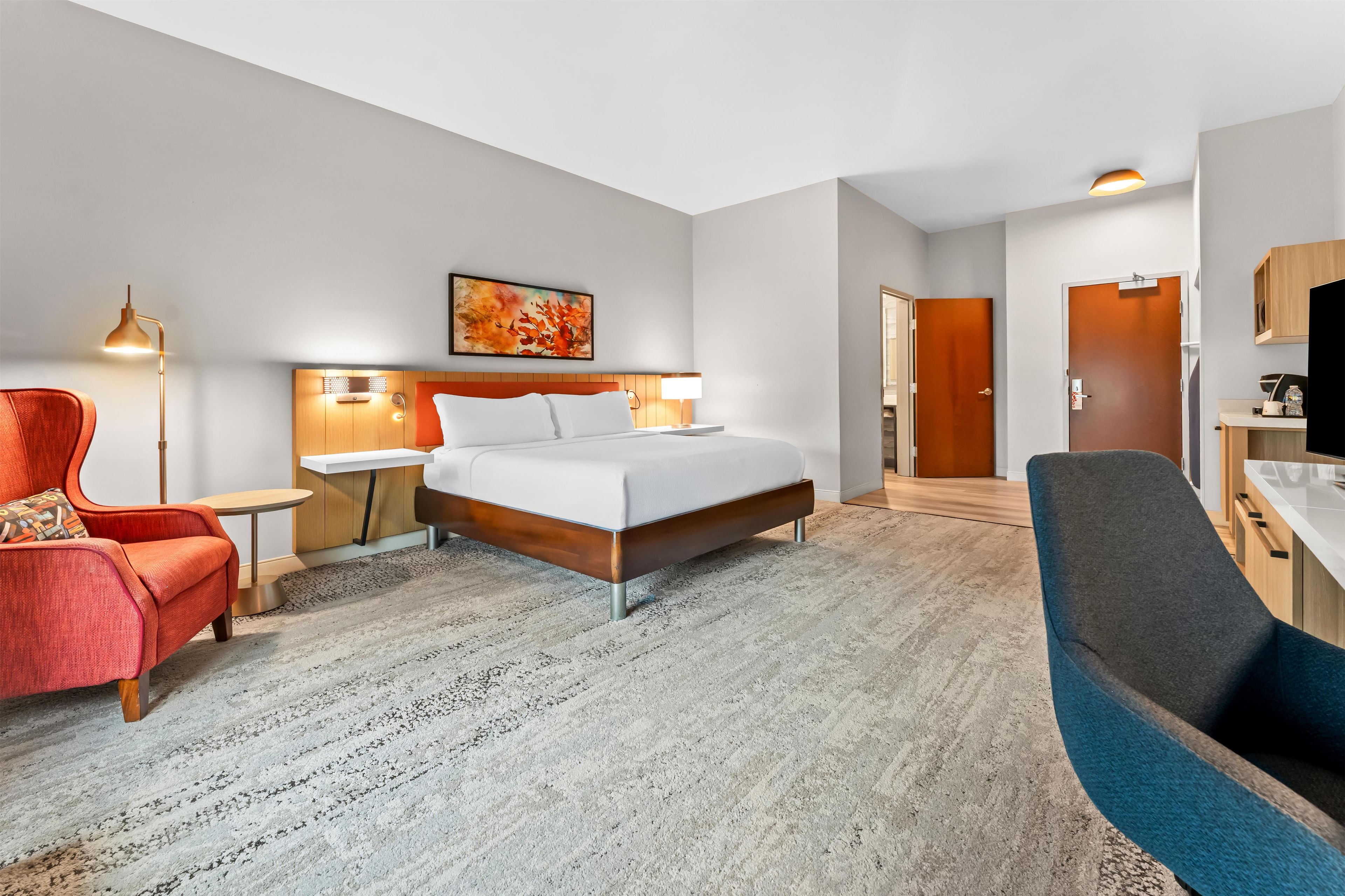 A hotel room with a bed, a desk, a chair, a lamp, and a painting on the wall.