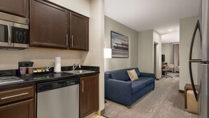 A modern kitchen with a built-in microwave, dishwasher, and a coffee maker. There is a blue sofa in the living area with a painting on the wall. - Homewood Suites Fort Wayne (Fort Wayne)