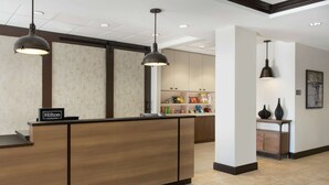 A hotel reception area with a wooden reception desk, two pendant lights, and a minibar stocked with snacks. - Homewood Suites Fort Wayne (Fort Wayne)