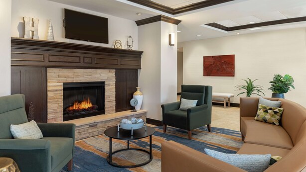 A modern living room with a fireplace, green and brown sofas, a coffee table, and decorative items. - Homewood Suites Fort Wayne (Fort Wayne)