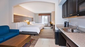A hotel room with a bed, a blue sofa, a small table, a kitchen area with a sink and cabinets, and a view of another room. - Holiday Inn Express & Suites Warminster - Horsham by IHG (Warminster)