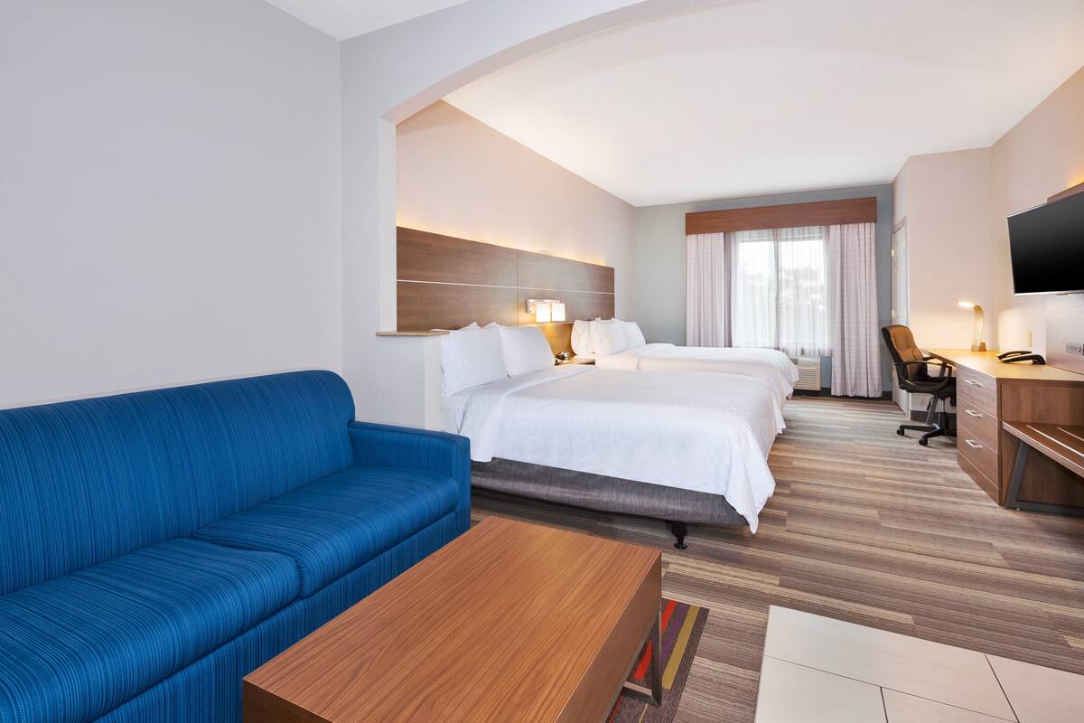A hotel room with a blue sofa, a wooden coffee table, a bed with white bedding, a desk with a chair, and a TV mounted on the wall.