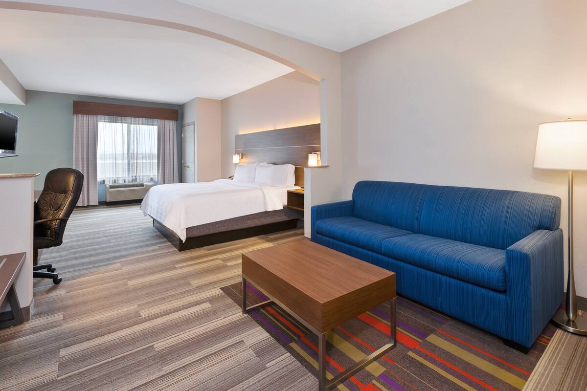 A hotel room with a bed, a blue sofa, a wooden coffee table, a desk with a chair, and a TV.