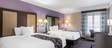 A hotel room with two beds, a desk, and a window with patterned curtains.