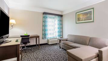 Camera d'albergo con scrivania, sedia, divano, televisore, lampada, condizionatore d'aria, tappeto decorato e finestra con tende.