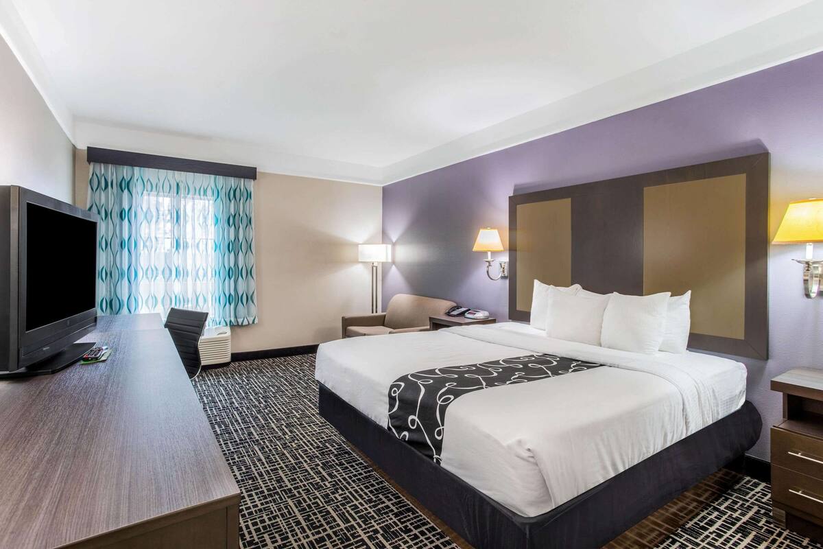 Ein Hotelzimmer mit einem großen Bett, einem Schreibtisch mit Fernseher, zwei Nachttischlampen und einem gemusterten Teppich.