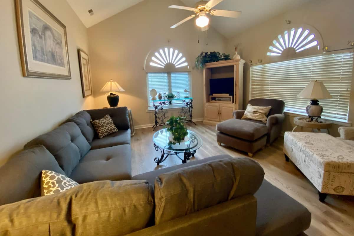 A spacious living room with a ceiling fan, a sofa, an ottoman, a coffee table, and a television cabinet.