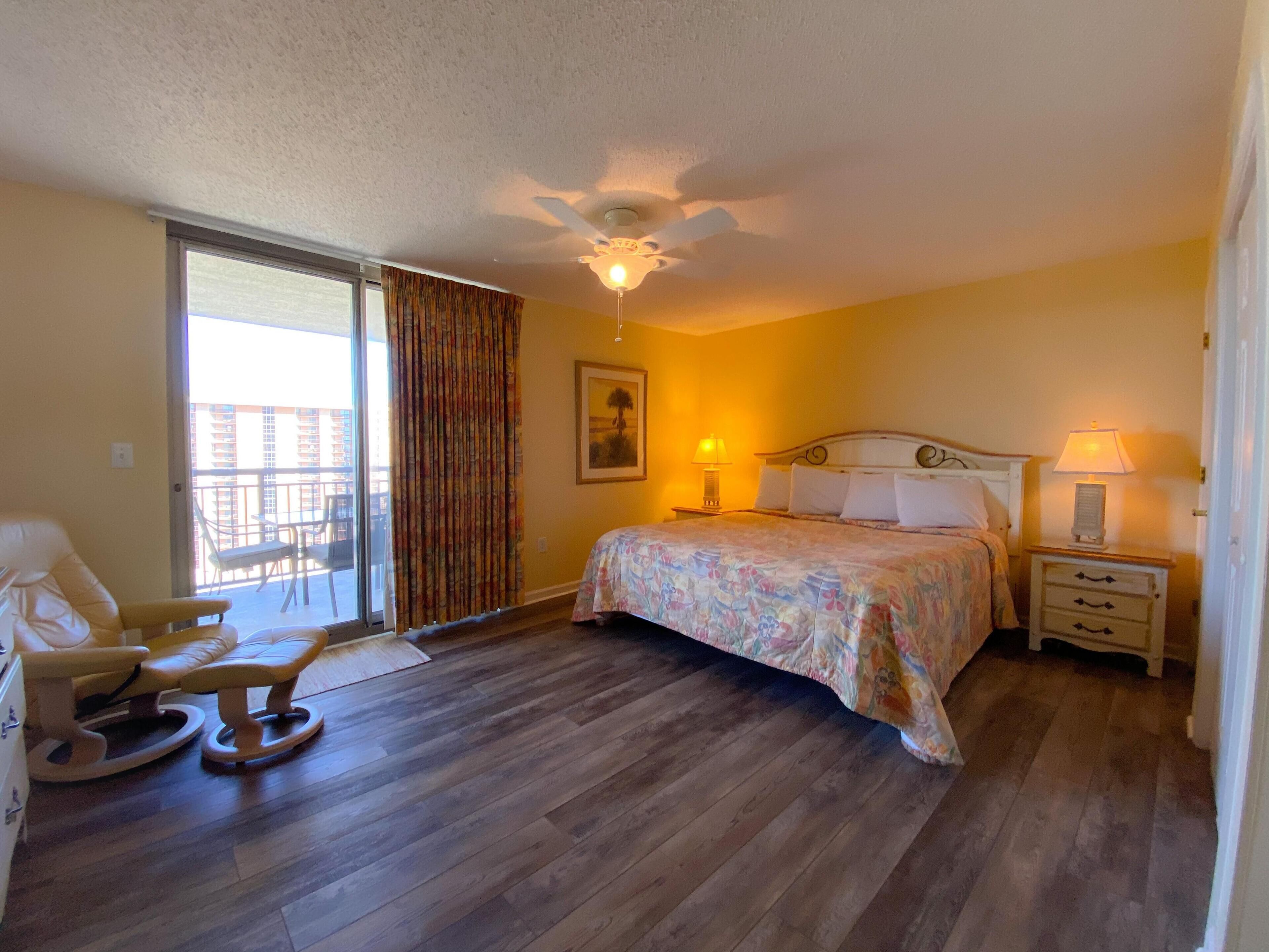 A bedroom with a bed, a chair, a nightstand, a lamp, a ceiling fan, and a window with curtains.