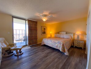 A bedroom with a bed, a chair, a nightstand, a lamp, a ceiling fan, and a window with curtains. - Palmetto Vacation Rentals at Kingston Plantation (Myrtle Beach)