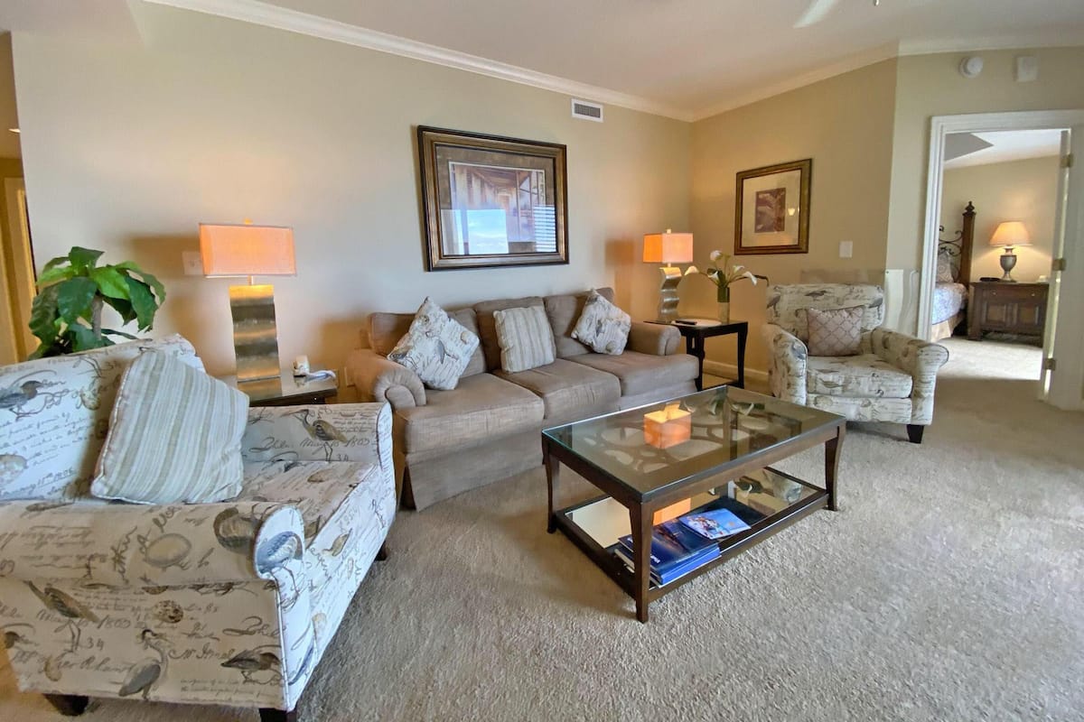 A living room with a beige carpet, two sofas, a glass coffee table, and two lamps.