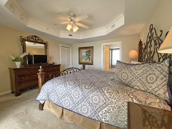 A bedroom with a bed, dresser, mirror, ceiling fan, and a door leading to another room.