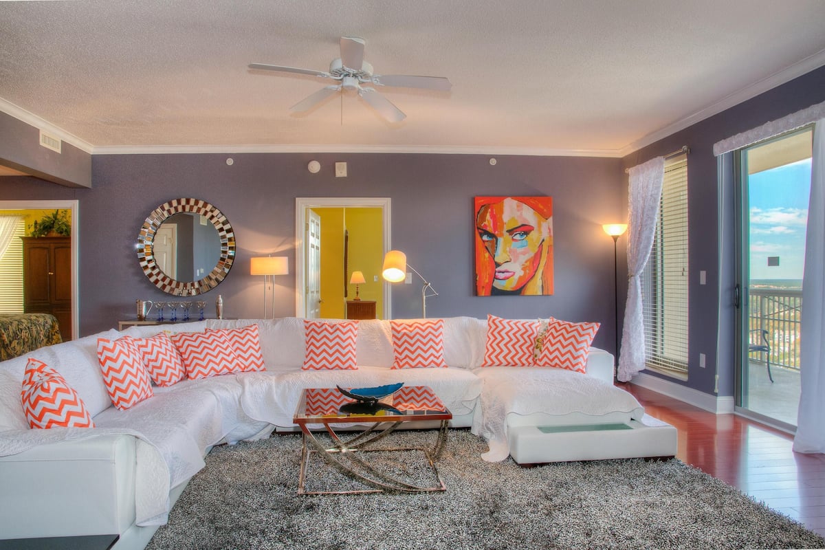 A living room with a white sectional sofa, a round mirror, a painting, and a ceiling fan.