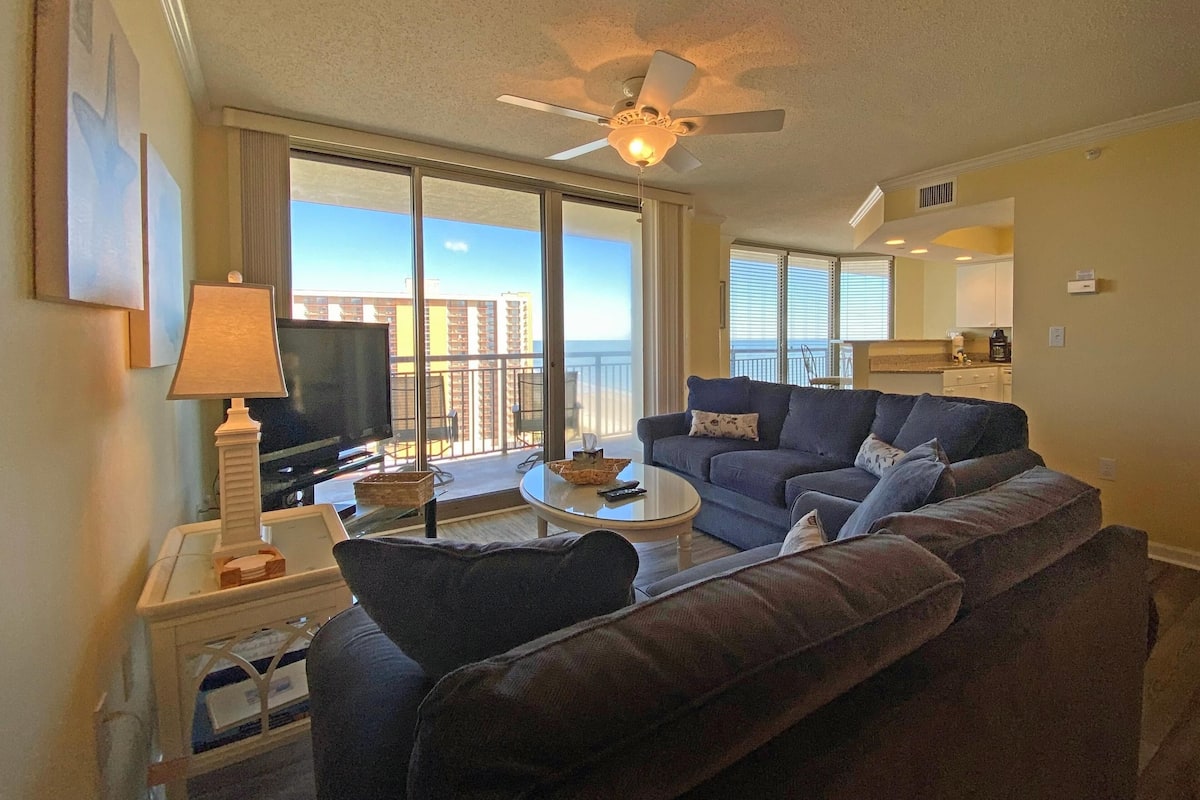 A living room with a sofa, a coffee table, a TV, and a ceiling fan. There is a balcony with a view of buildings.