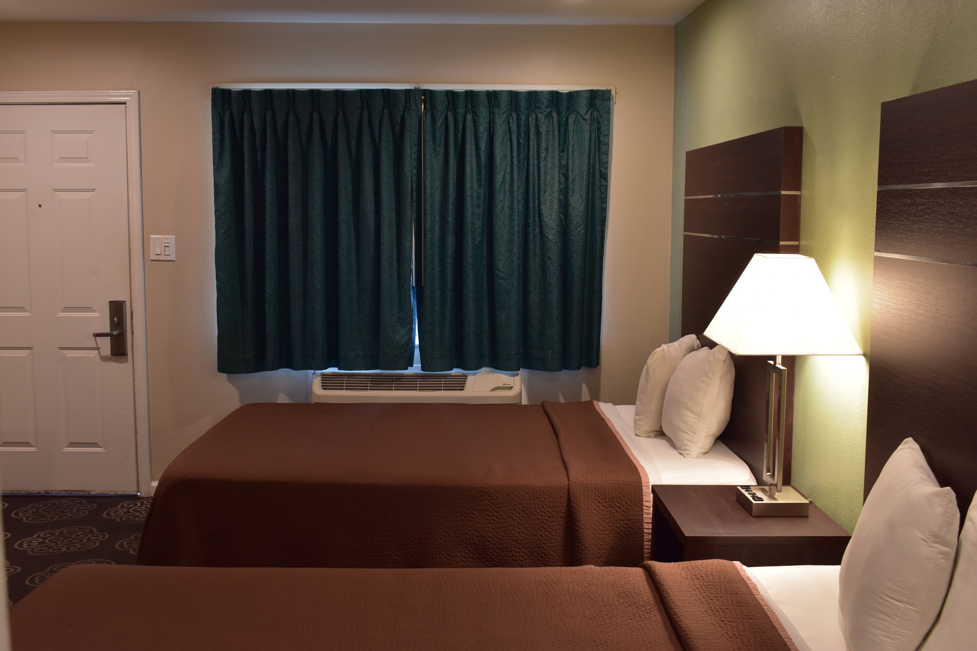 Ett hotellrum med två sängar, ett fönster med gardiner, en sänglampa och en sänggavel som är monterad på väggen.