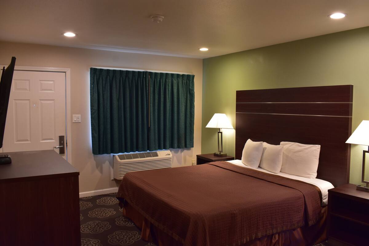 A hotel room with a bed, a desk, a lamp, a window with curtains, and an air conditioning unit.