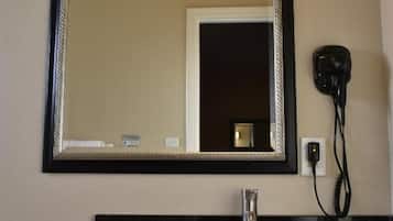 Casa de banho com bancada preta, lavatório branco, espelho com moldura preta e um secador de cabelo ligado.