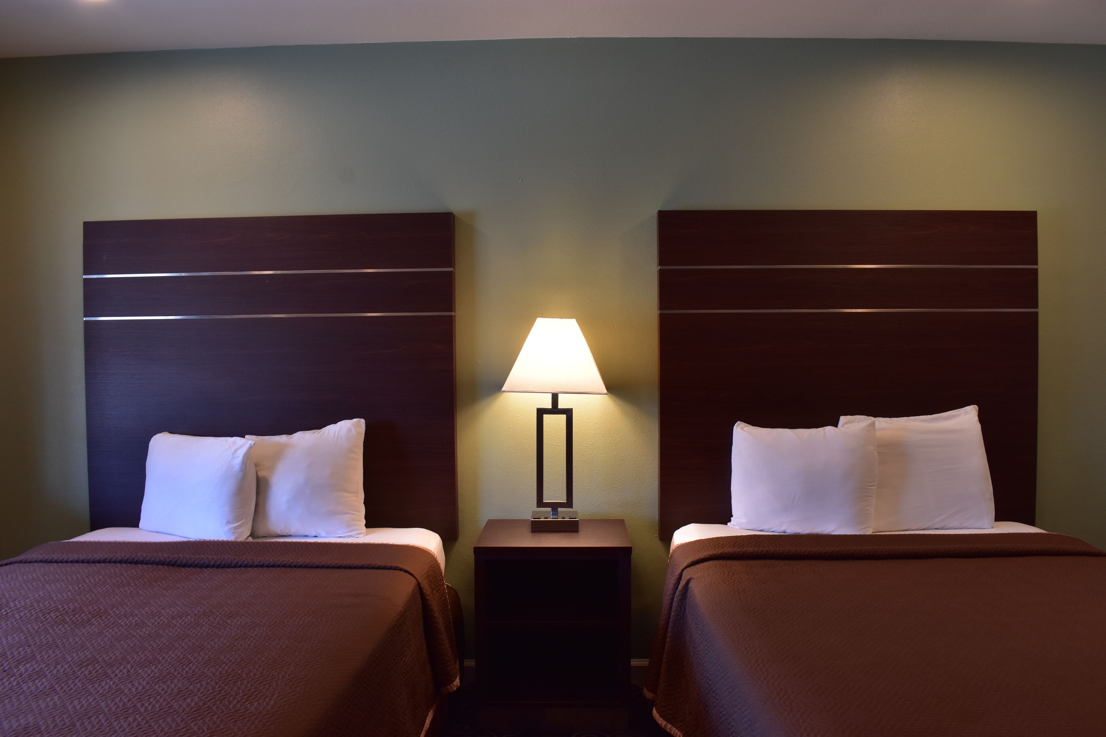 A hotel room with two beds, a nightstand with a lamp, and a green wall.