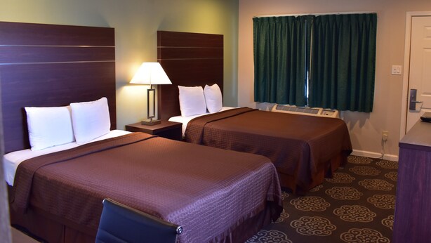 Quarto de hotel com duas camas, mesa de cabeceira com abajur, janela com cortinas e carpete estampado.