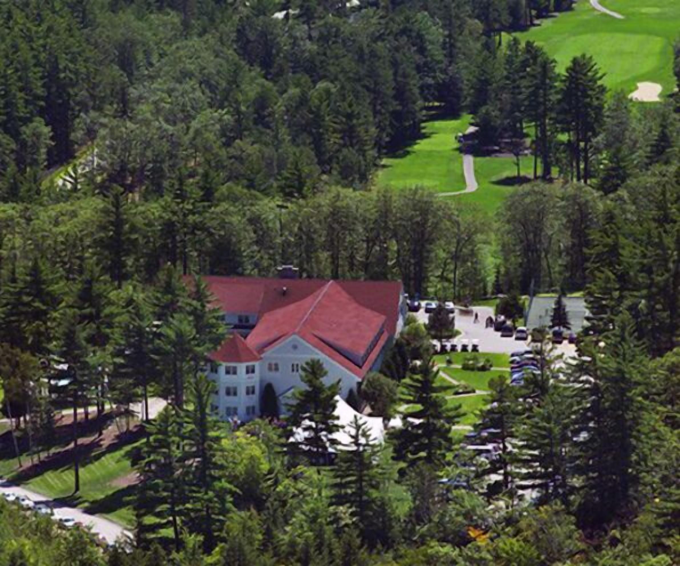 A large building with a red roof surrounded by dense forest and a golf course.