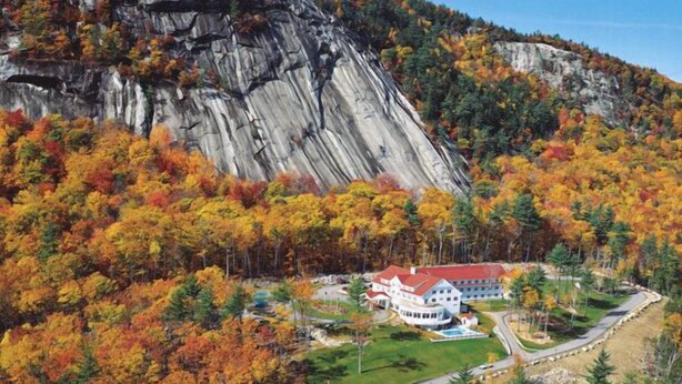 Un hotel con techo rojo, rodeado de follaje otoñal y montañas rocosas al fondo.