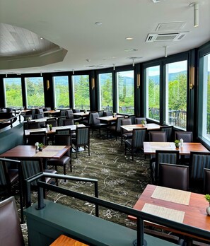 Een restaurant met meerdere tafels en stoelen, grote ramen met uitzicht op groen en een plafond met inbouwverlichting.