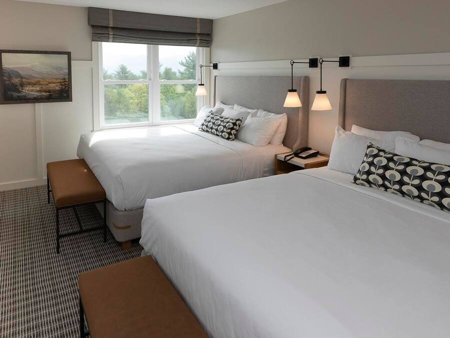 A hotel room with two beds, a bench, a nightstand, and a window with a view of trees.