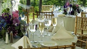 Un montaje para comer al aire libre con mesas preparadas para una comida, rodeado de sillas y decorado con flores.