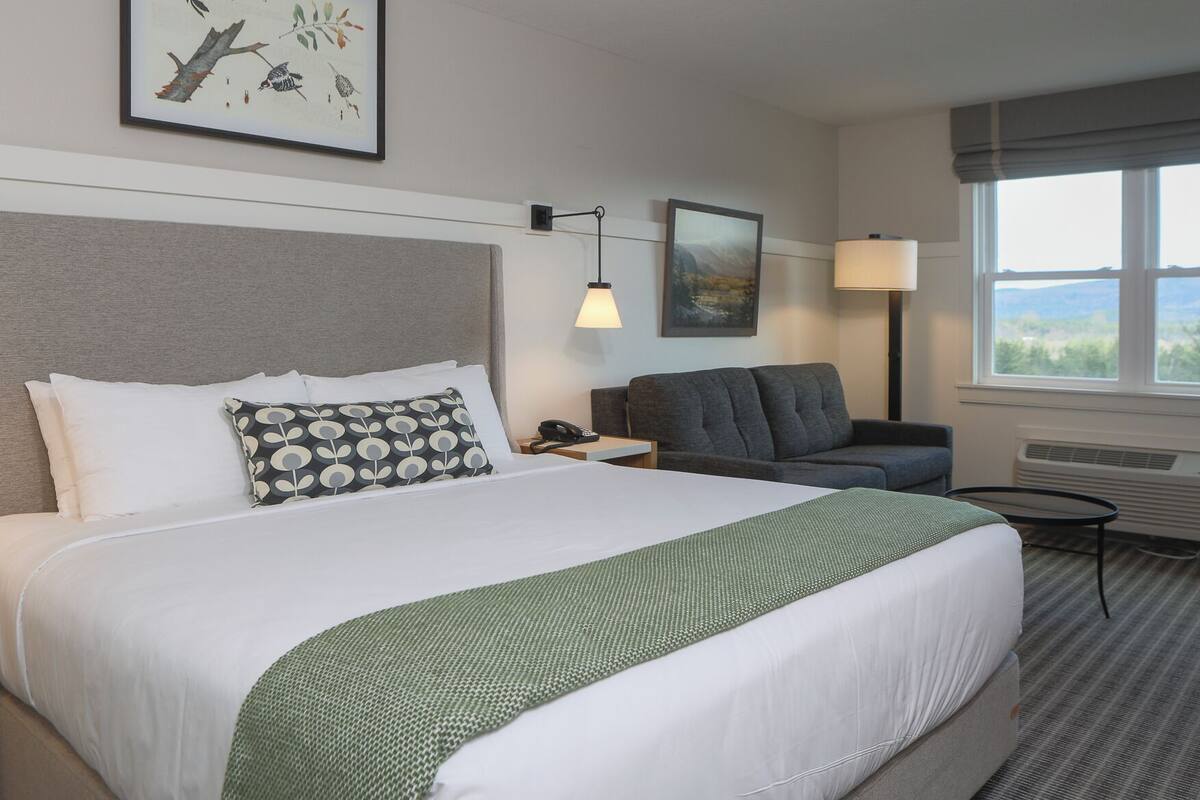 Une chambre d’hôtel avec un lit, une chaise, un bureau avec une lampe et une fenêtre donnant sur un paysage agréable.