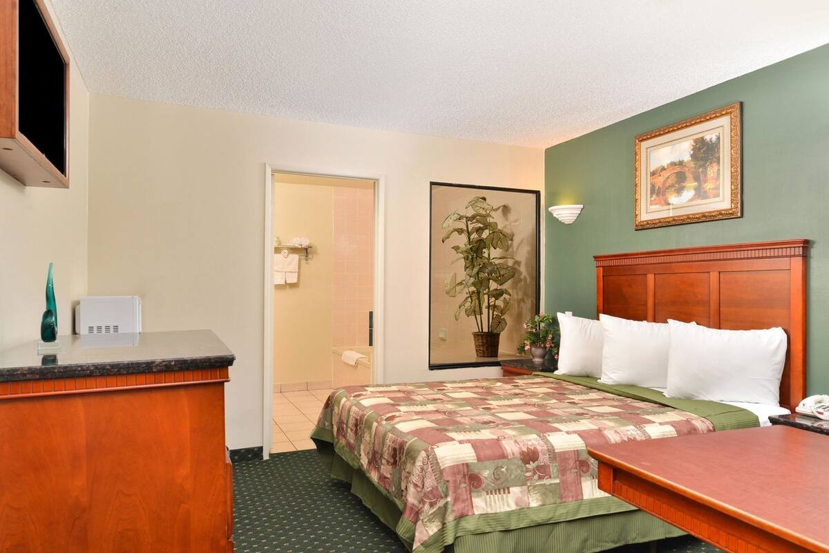 A hotel room with a bed, a desk, a telephone, a painting, and a potted plant.