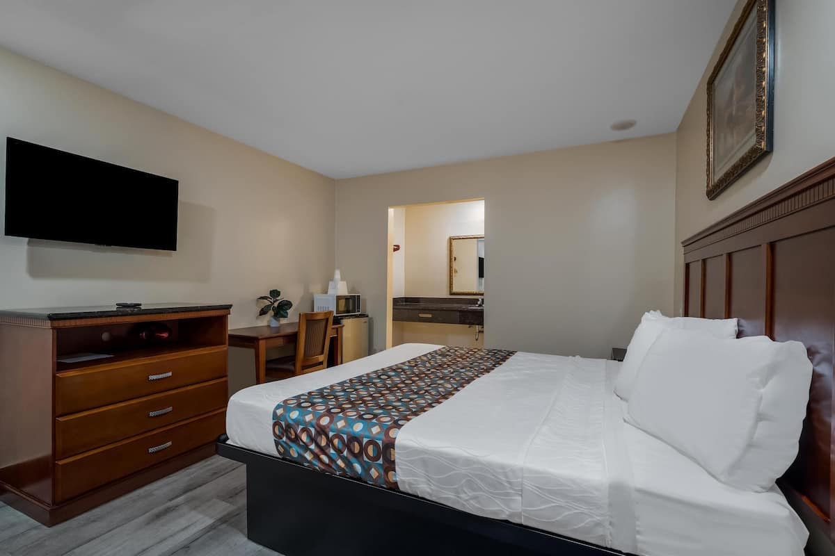 A hotel room with a bed, a desk, a chair, a flat-screen TV, a mirror, and a wooden dresser.
