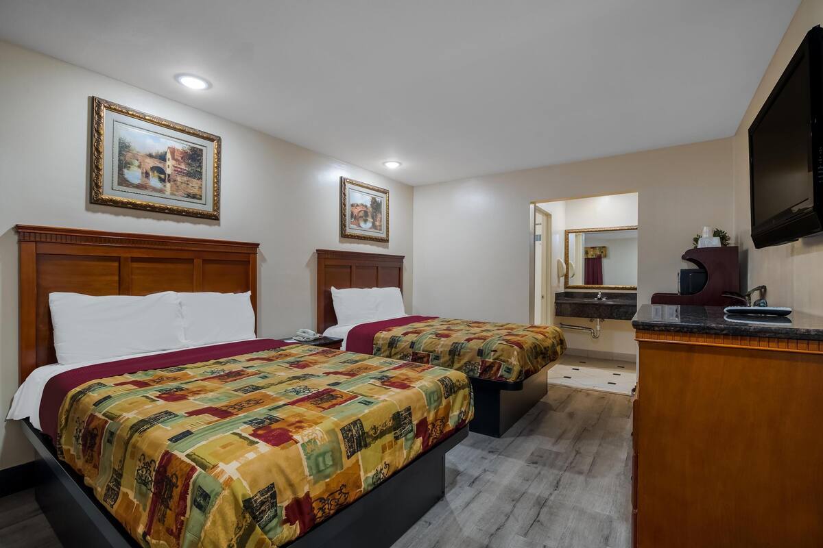 A hotel room with two beds, a wooden headboard, a large mirror, a TV, and a small desk with a sink.