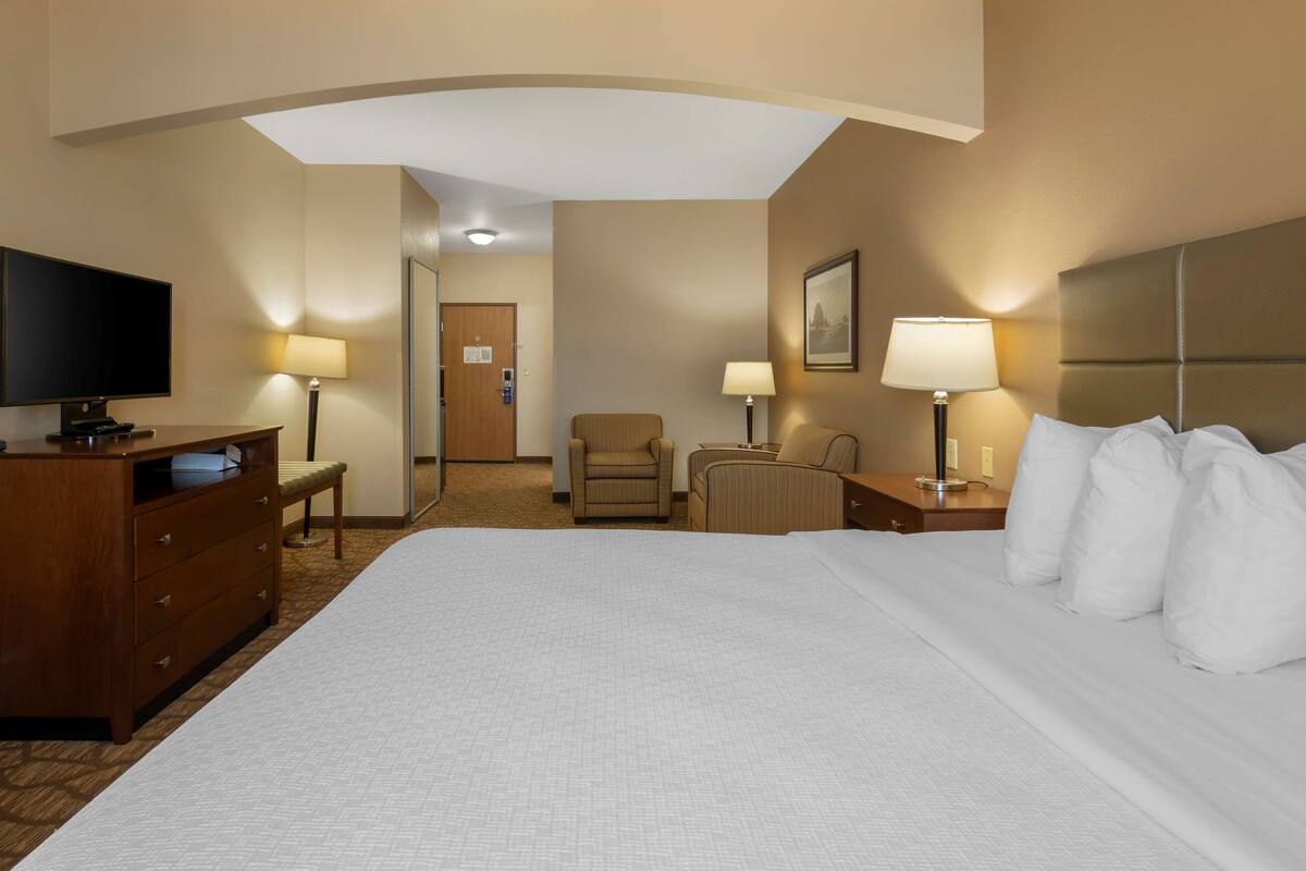 A hotel room with a large bed, a desk with a flat-screen TV, two chairs, a nightstand with a lamp, and a door leading to a hallway.
