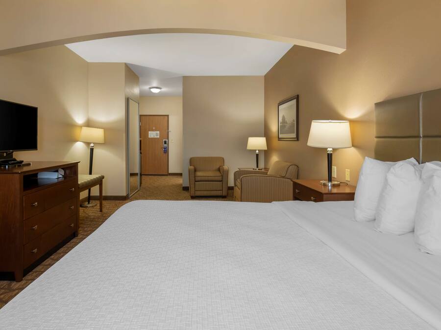 Habitación de hotel con una cama grande, un escritorio con televisor de pantalla plana, dos sillas, una mesita de noche con lámpara y una puerta que da a un pasillo.