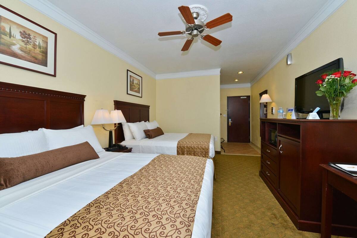 Ein Hotelzimmer mit zwei Betten, einem Holz-Kopfteil, einem Deckenventilator, einem Fernseher, einem Schreibtisch und einer Vase mit Blumen.