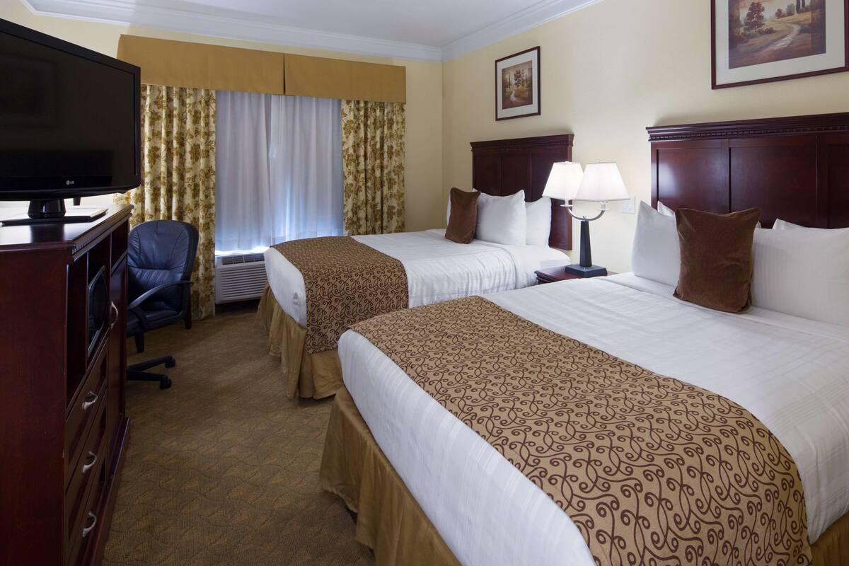 Ein Hotelzimmer mit zwei Betten, einem Schreibtisch mit Fernseher, einem Sessel und einem Fenster mit Vorhängen.